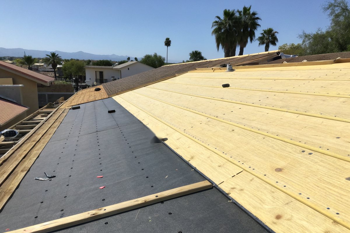Fresh plywood roof decking with gray synthetic underlayment being fastened down in overlapping courses on a sunny day