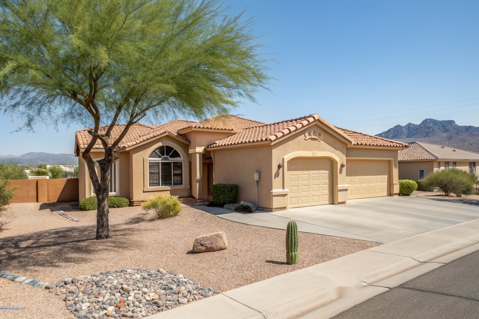 Tan stucco home with a terracotta tile roof and arched window behind a palo verde tree in a gravel yard, desert mountains on the horizon under a clear morning sky