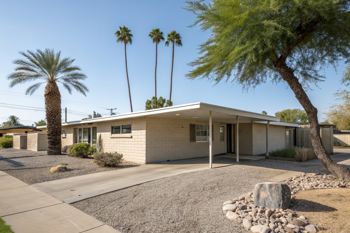 1960s cream slump-block ranch home with a low, nearly flat roof and a carport, palms and a palo verde in a gravel yard
